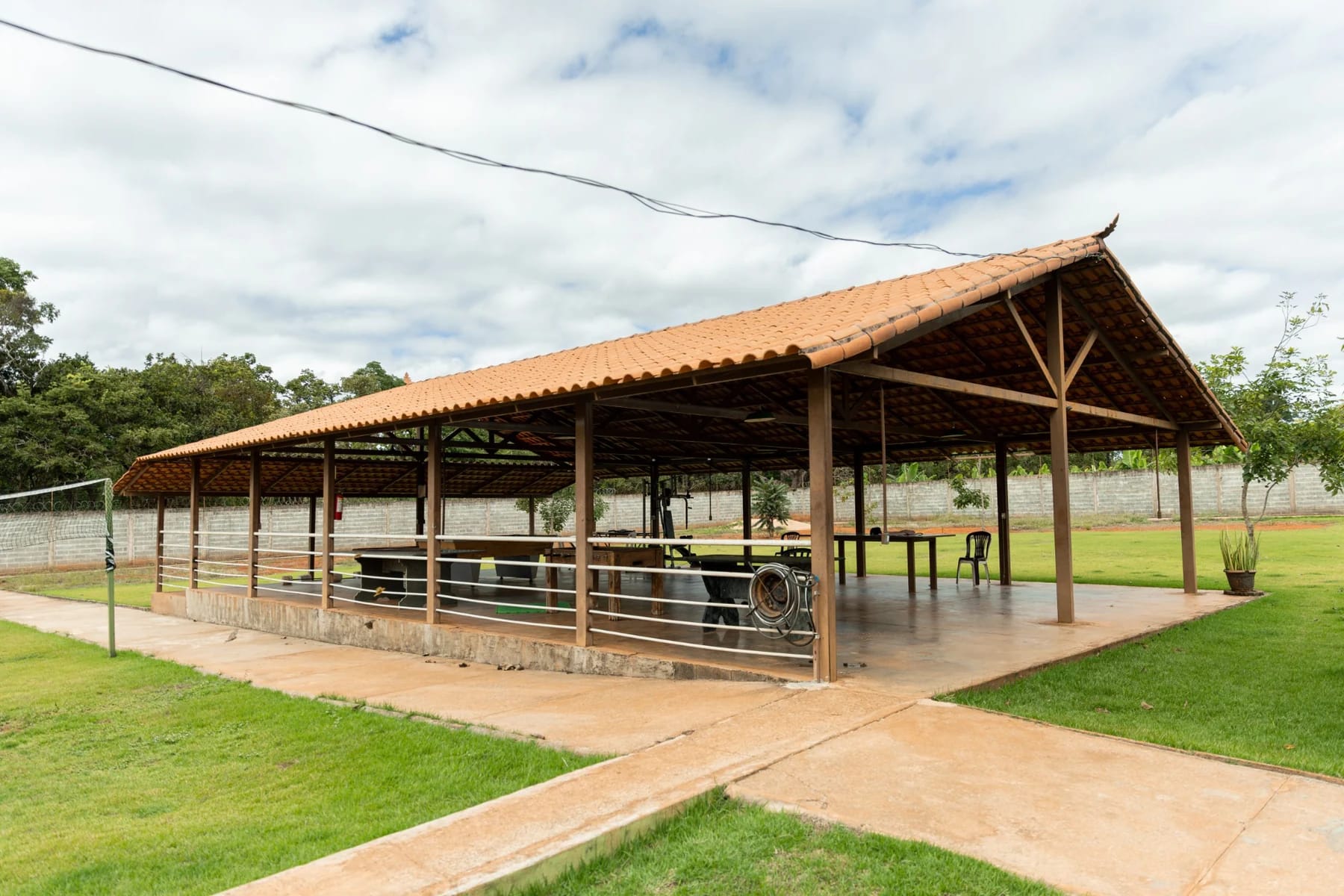 Pavilhão aberto com cobertura de telhas, mesas e área gramada ao redor.