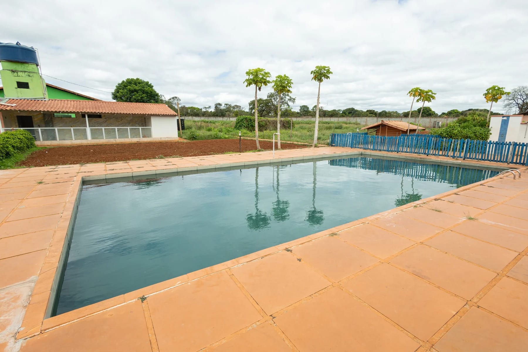 Piscina externa com circulação pavimentada, vegetação e construções ao redor.